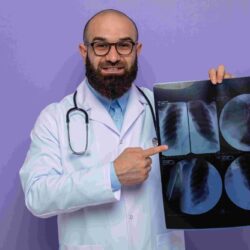 compressed_bearded-man-doctor-white-coat-with-stethoscope-around-neck-wearing-glasses-holding-x-ray-pointing-with-index-finger-it-smiling-cheerfully-standing-purple-background (1)