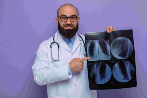 compressed_bearded-man-doctor-white-coat-with-stethoscope-around-neck-wearing-glasses-holding-x-ray-pointing-with-index-finger-it-smiling-cheerfully-standing-purple-background (1) compressed_bearded-man-doctor-white-coat-with-stethoscope-around-neck-wearing-glasses-holding-x-ray-pointing-with-index-finger-it-smiling-cheerfully-standing-purple-background (1)