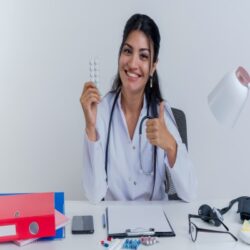 happy-young-female-doctor-wearing-medical-robe-stethoscope-sitting-desk-with-medical-tools-holding-pack-medical-tablets-looking-showing-thumb-up-isolated (4) (1)
